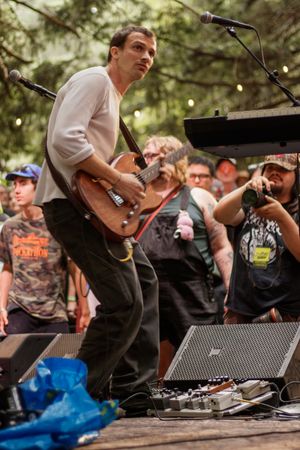 Guitarist in white shirt performs on outdoor stage with crowd watching and guitar pedals at his feet.