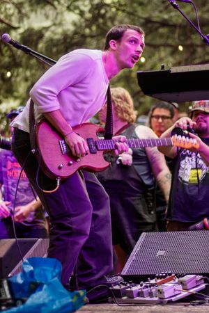 Guitarist performing on outdoor stage with pink electric guitar, surrounded by crowd and effects pedals.
