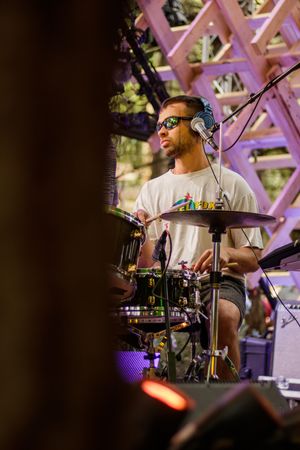 A drummer wearing sunglasses and headphones performs on stage outdoors at a live music event.