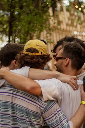 Group of friends hugging outdoors at a festival, one wearing a yellow cap, surrounded by trees.