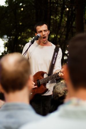 Passionate male vocalist in white tee sings into microphone while playing brown electric guitar outdoors.