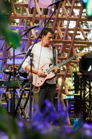 Musician playing a pink electric guitar on stage with wooden lattice backdrop and purple lighting.