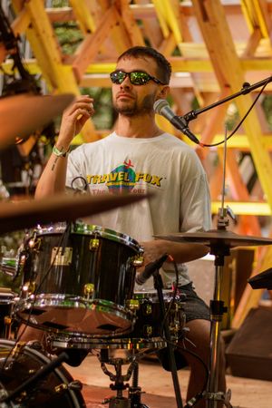 Drummer wearing sunglasses and Travel Fox t-shirt plays drums at indoor music venue with yellow scaffolding.