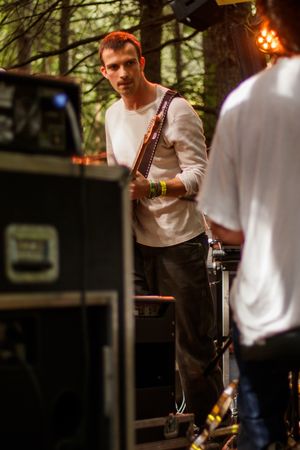 Musician with guitar strap stands backstage at outdoor forest music festival in white long sleeve shirt.