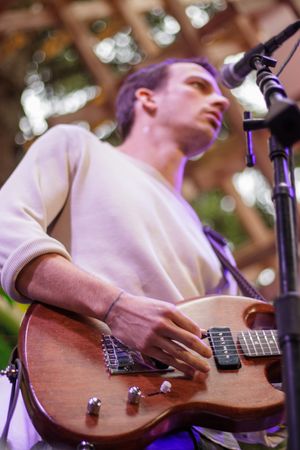 Musician playing a brown electric guitar on stage under purple lights with a microphone stand in front.
