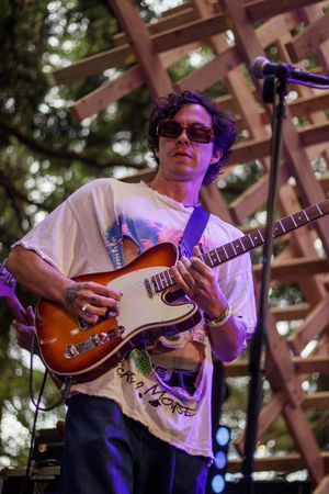 Young male guitarist in sunglasses plays a sunburst Telecaster on an outdoor stage with wooden lattice backdrop.