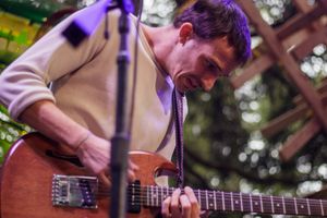 Musician playing electric guitar on outdoor stage, focused expression, wearing cream sweater at live concert.