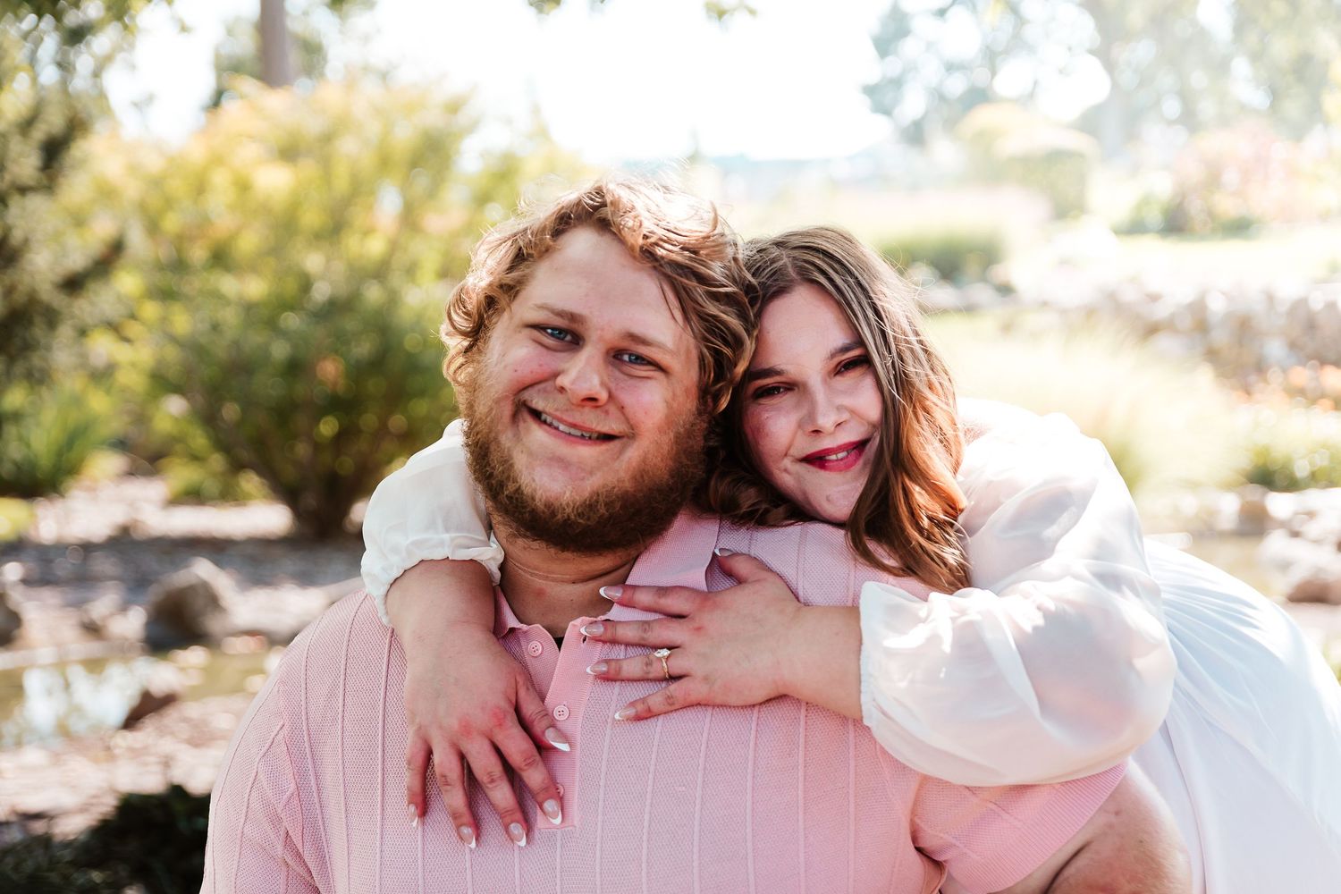 Happy couple posing outdoors, woman hugging man from behind, both smiling, wearing pink and white outfits.