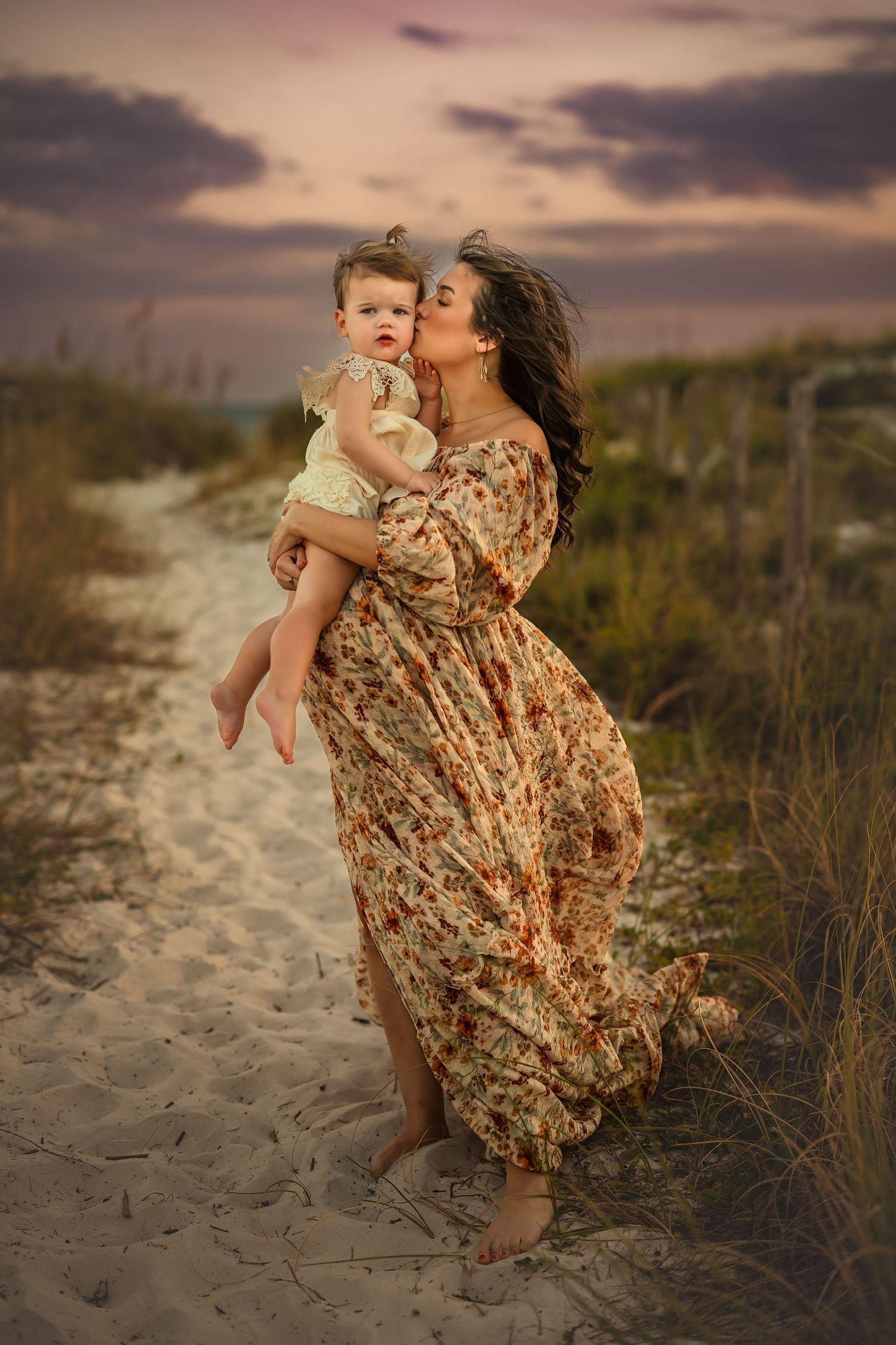 A woman in a floral dress kisses her baby on a sandy beach path at sunset.