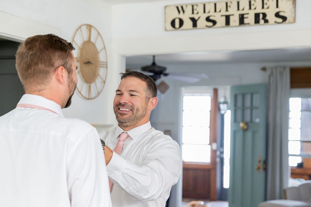 Two men in white dress shirts smiling indoors near a Wellfleet Oysters sign during a wedding preparation.