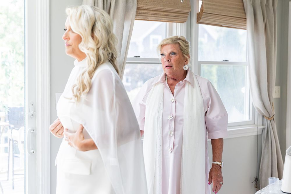 Two elegant women in white outfits standing near bright windows in a beautifully decorated room.