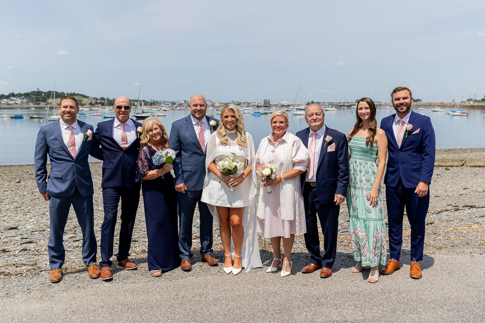 Wedding party group photo posing outdoors near a harbor on a sunny day, wearing formal attire.