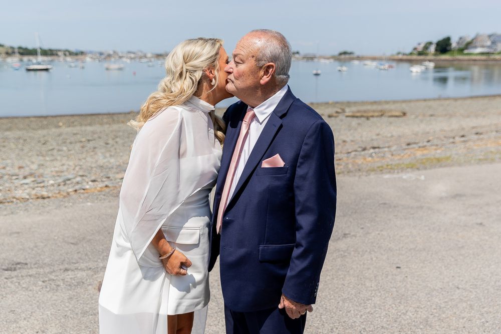 Couple sharing a romantic kiss on a beach, man in navy suit with pink tie, woman in white dress, harbor in background.