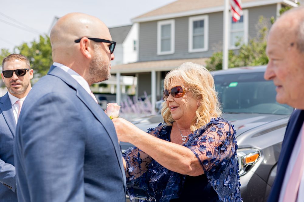 People greeting each other outdoors near cars, wearing sunglasses and formal attire on a sunny day.