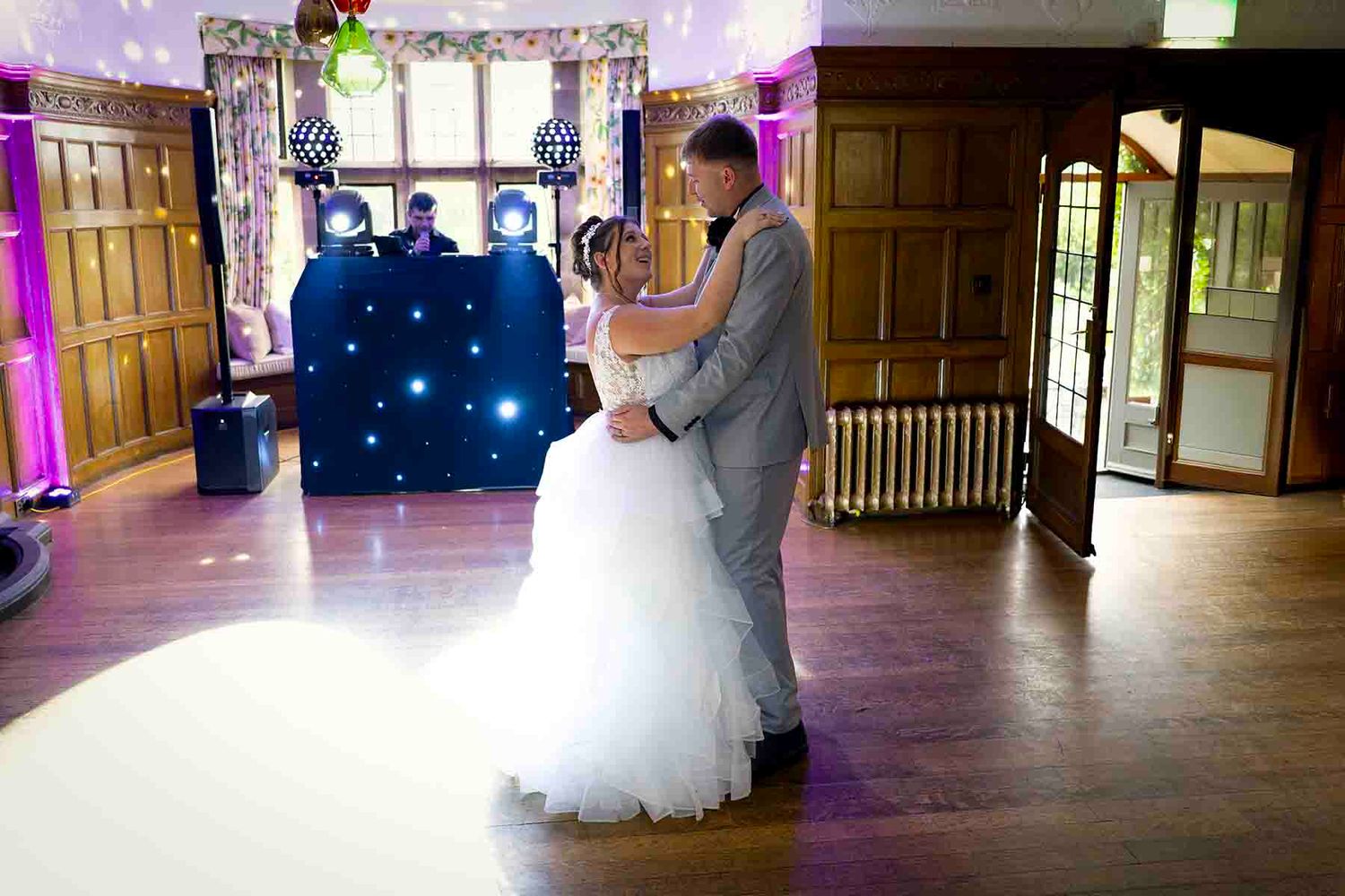 Bride and groom share their first dance in an elegant wedding venue with purple lighting and a DJ booth.