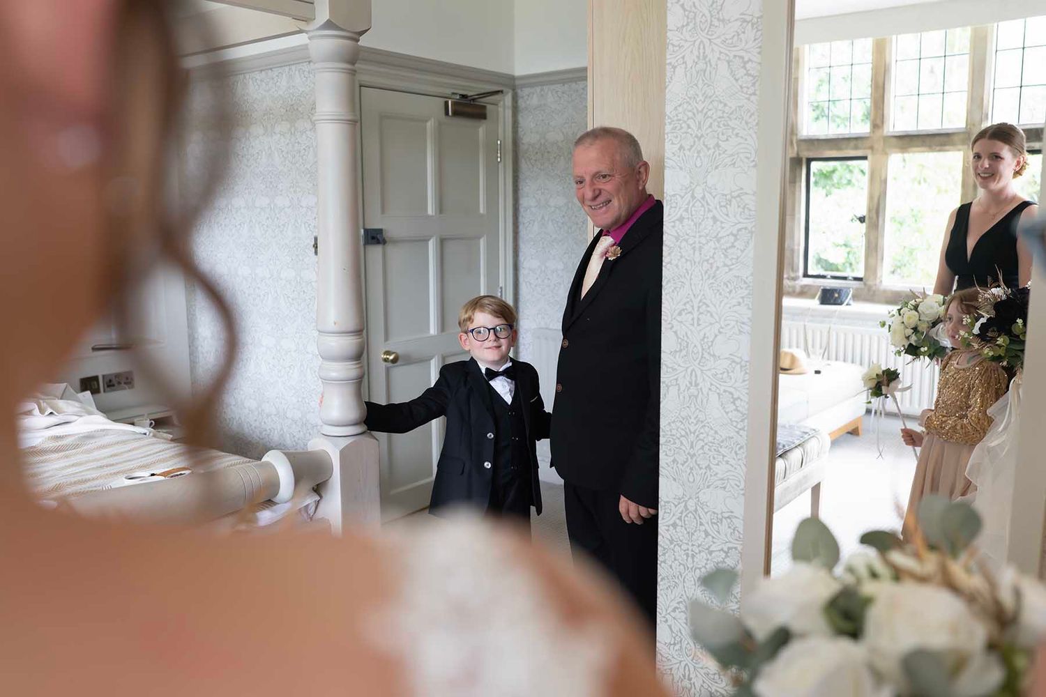 A young boy in a suit and an older man in a tuxedo smile at a bride during a wedding ceremony.