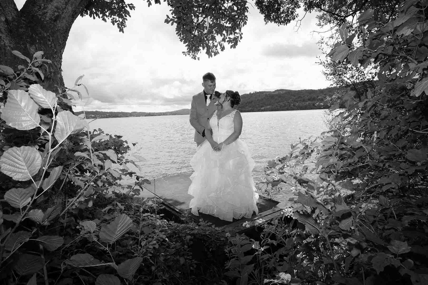 Black and white wedding photo of bride and groom embracing by a lakeside, framed by lush foliage and trees.