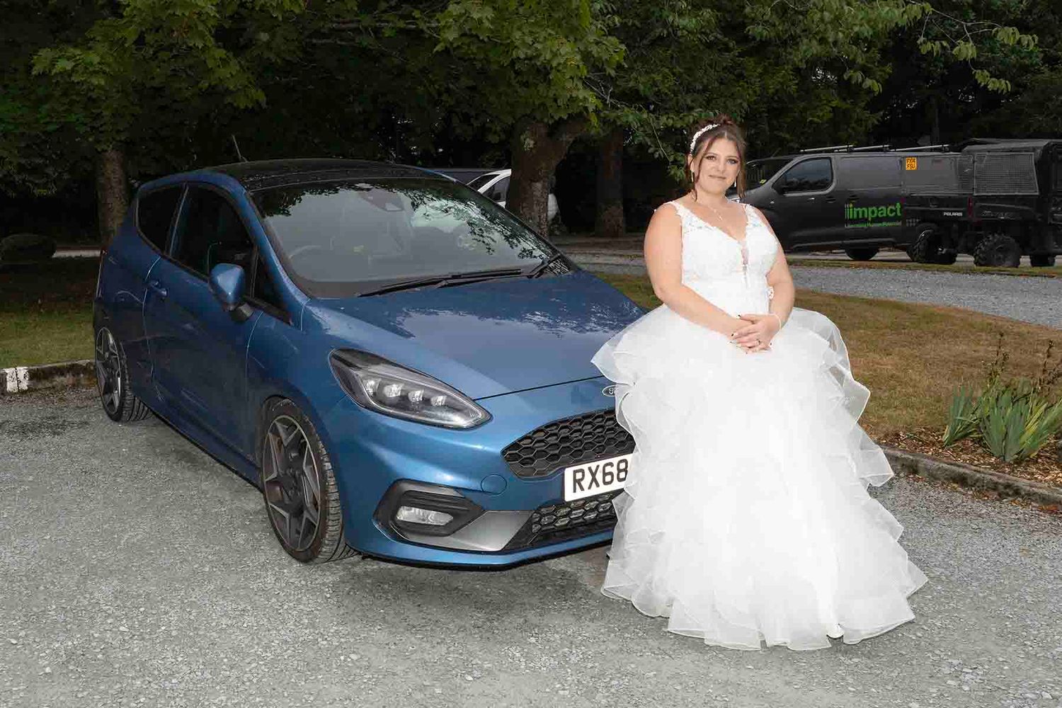 Bride in white wedding gown posing beside a blue Ford Fiesta ST on a gravel driveway surrounded by trees.