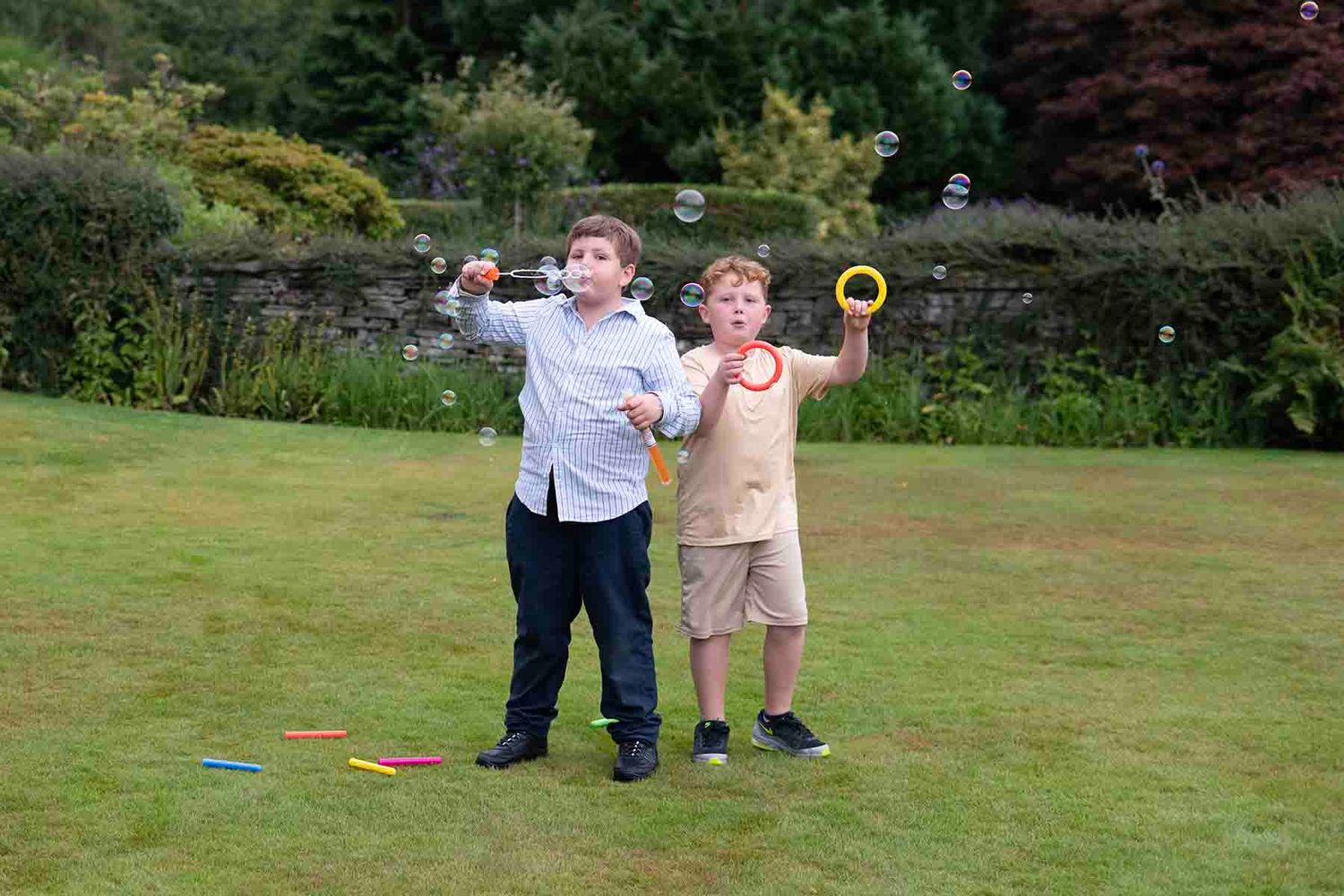 Two boys blowing bubbles in a lush green garden on a sunny day.