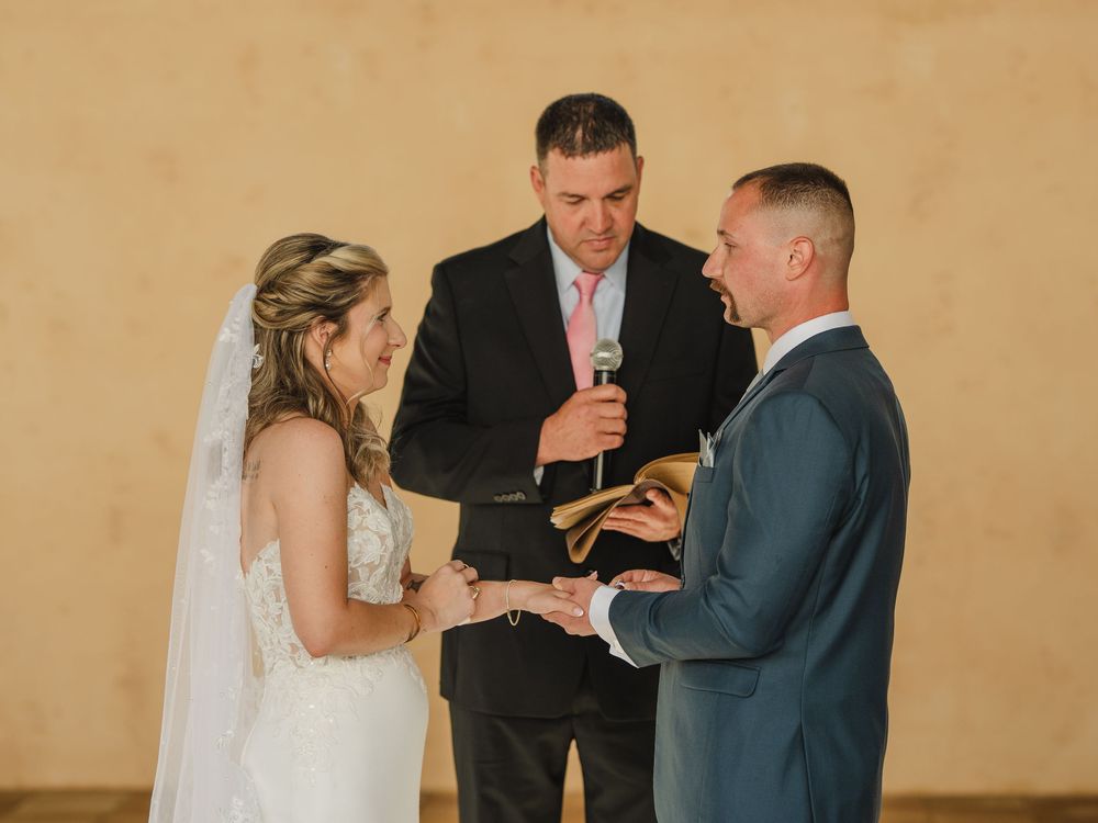 Bride and groom exchange vows at wedding ceremony with officiant holding book outdoors.