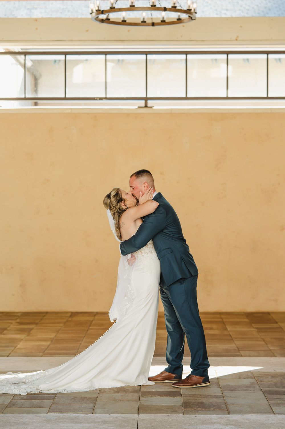 Bride and groom share a romantic dip kiss in a sunlit venue with warm beige walls and elegant chandelier.