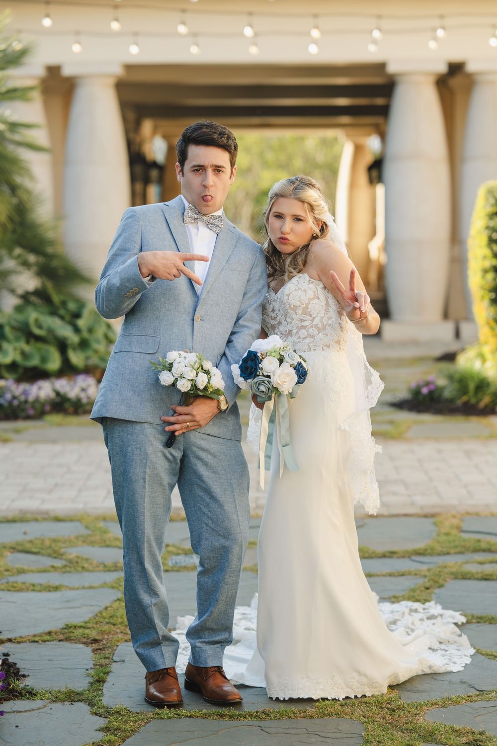 Bride in lace gown and groom in light blue suit pose playfully outdoors at elegant wedding venue with floral bouquets.
