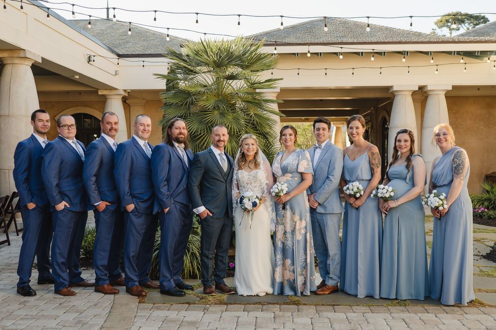 Wedding party posing outdoors in blue suits and dusty blue bridesmaid gowns at an elegant venue with string lights.