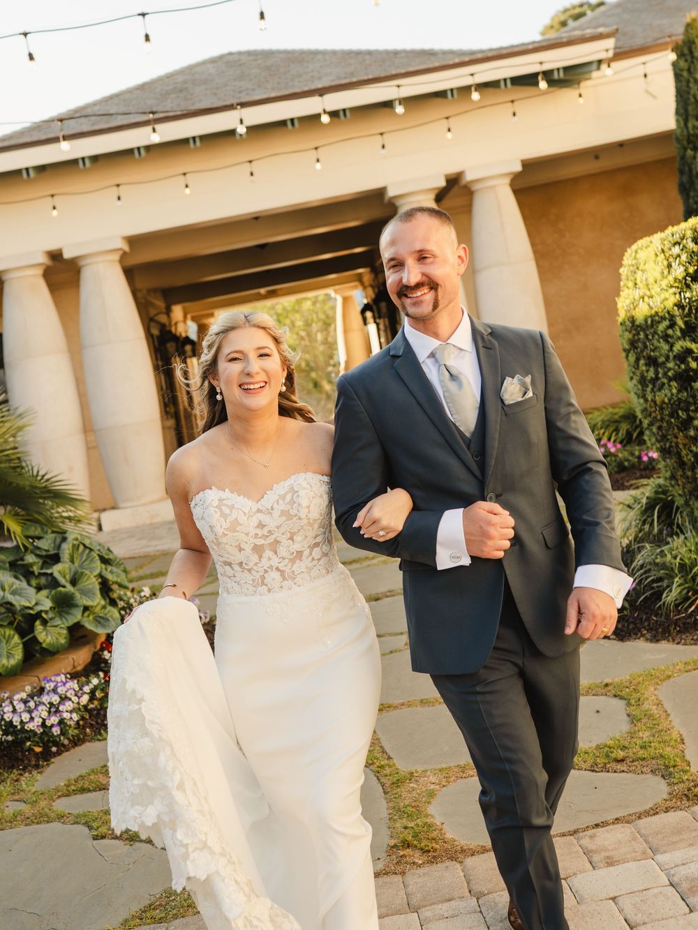 A joyful bride in a lace gown and groom in a navy suit walk hand-in-hand outside an elegant venue.