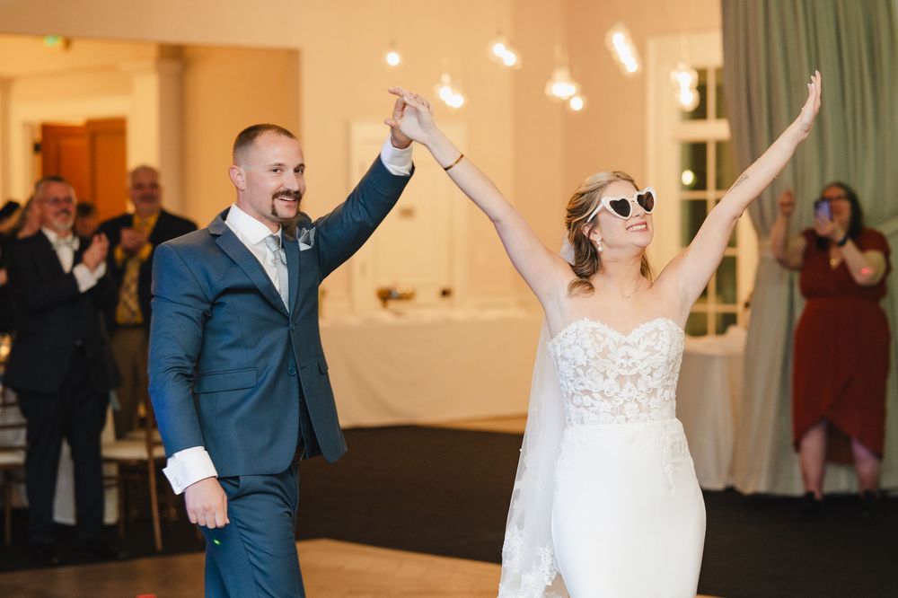 Joyful bride in white gown and sunglasses raises arms with groom in blue suit during wedding reception grand entrance.