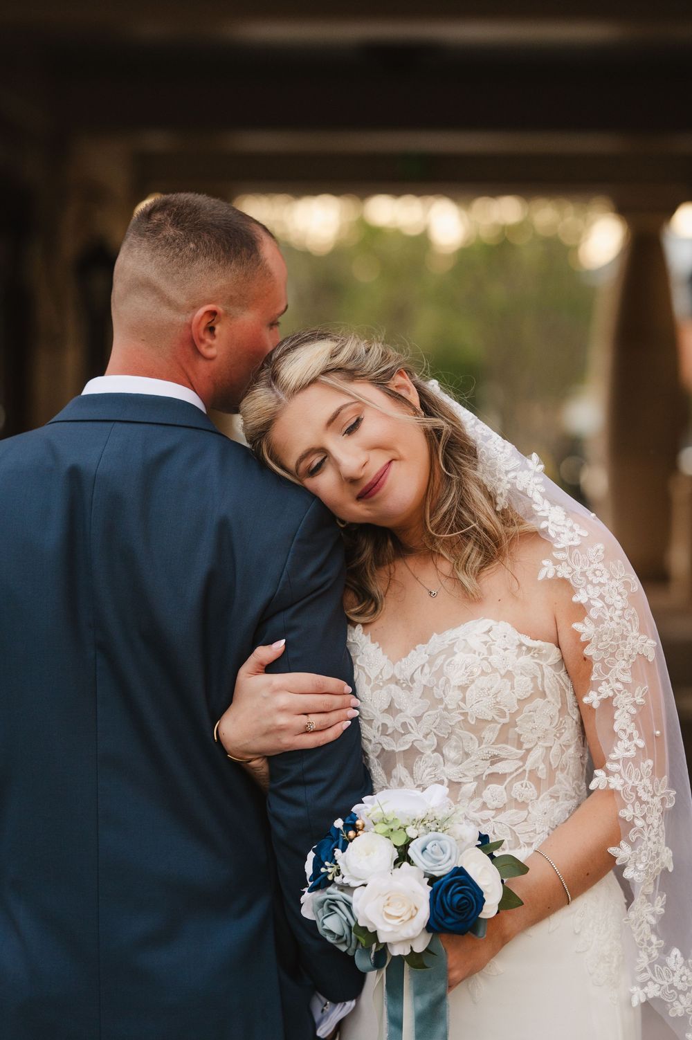Bride in lace gown holding blue and white bouquet leans on groom in navy suit during romantic wedding portrait.