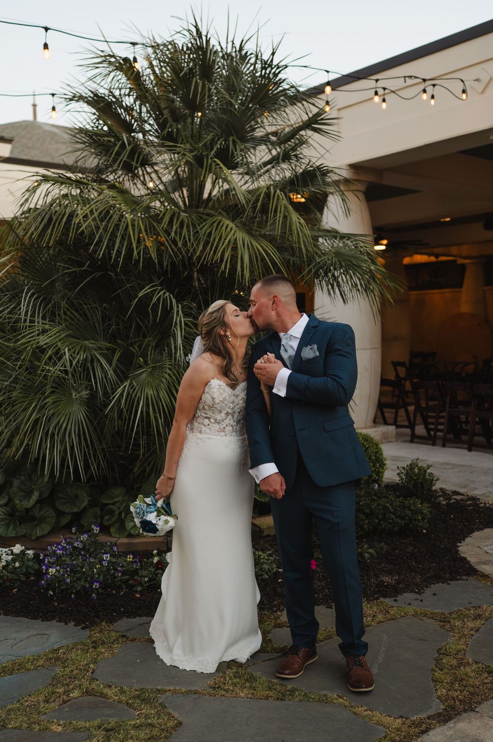 Bride and groom share a romantic kiss outdoors at dusk, surrounded by lush palm trees and string lights at their wedding venue.