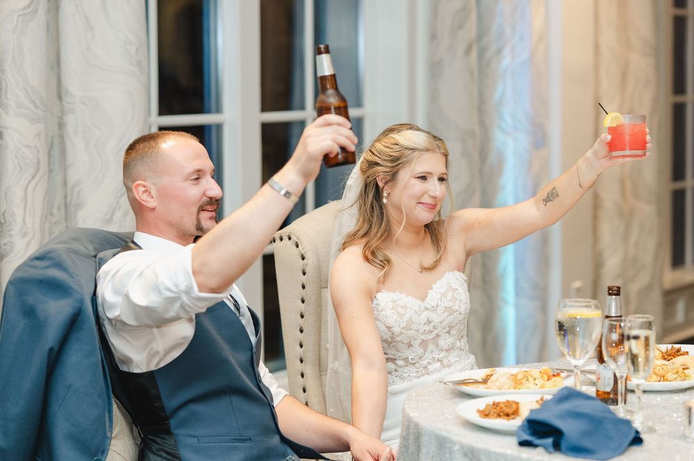 Bride and groom raising glasses in a toast at their wedding reception, smiling joyfully at a decorated table.