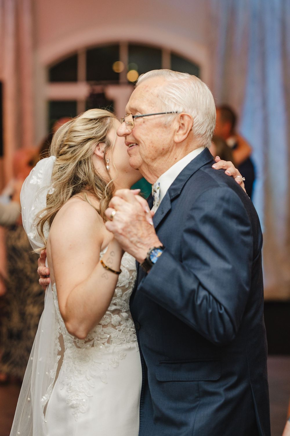 Bride and elderly man share a tender dance and kiss at a wedding reception in an elegant venue.