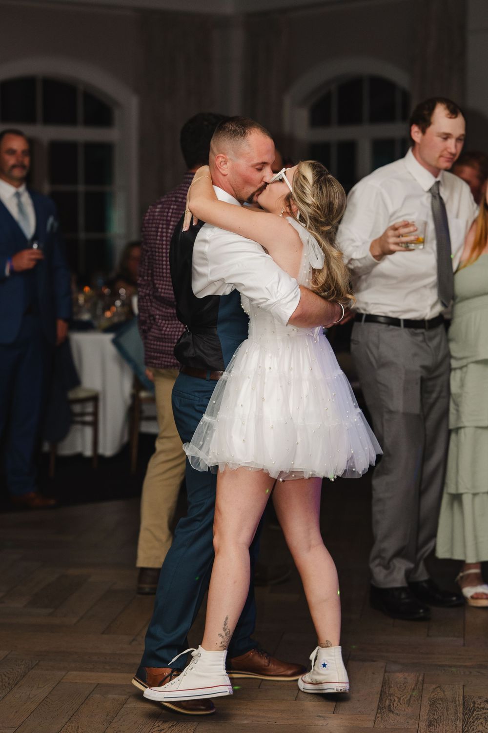Bride in short white tulle dress and Converse sneakers shares first dance kiss with groom at elegant wedding reception.