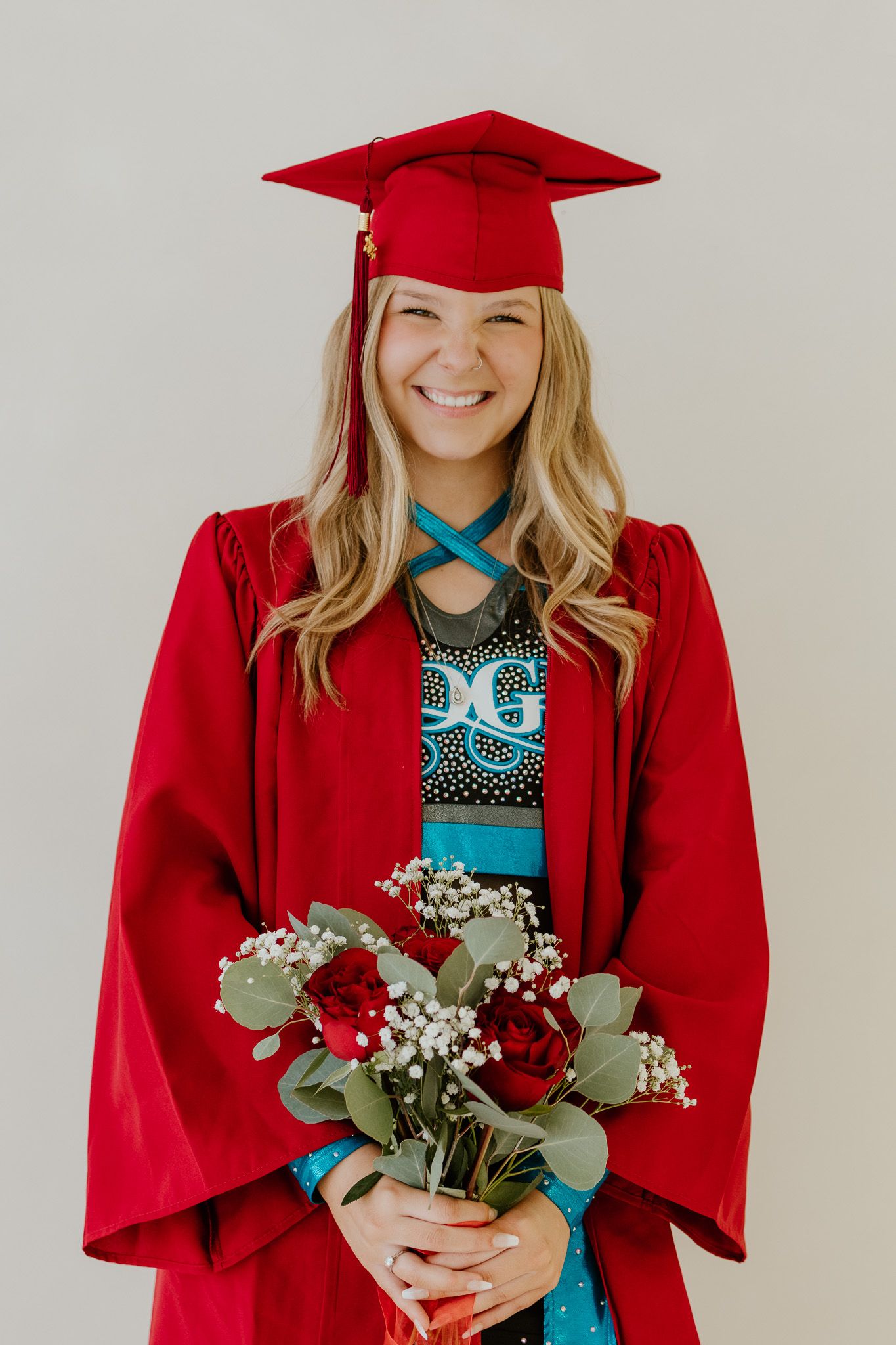 Smiling graduate in red cap and gown holding red roses and baby's breath bouquet against neutral background.