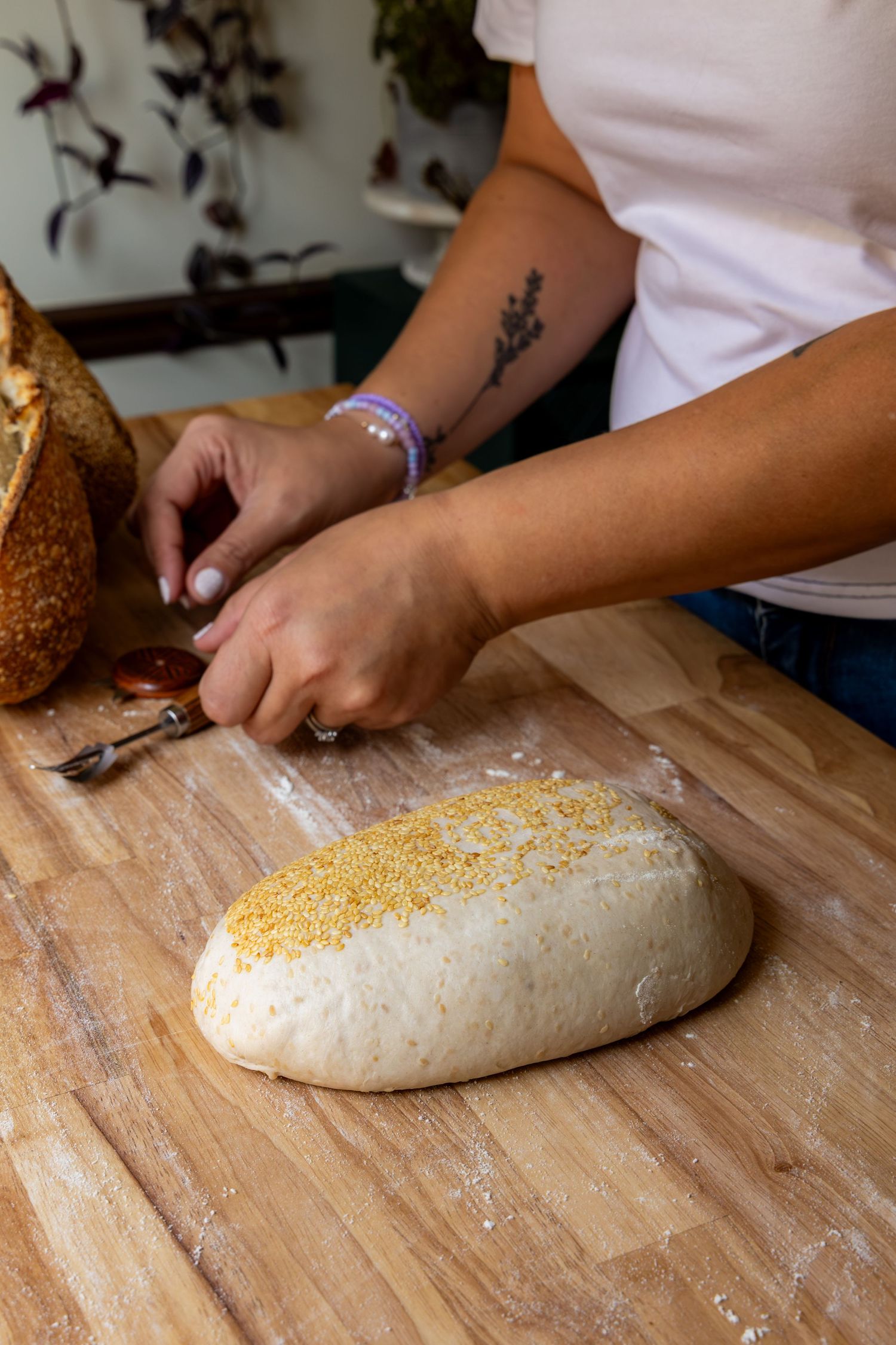 Baker scoring bread dough topped with cornmeal on a wooden cutting board in a rustic kitchen.