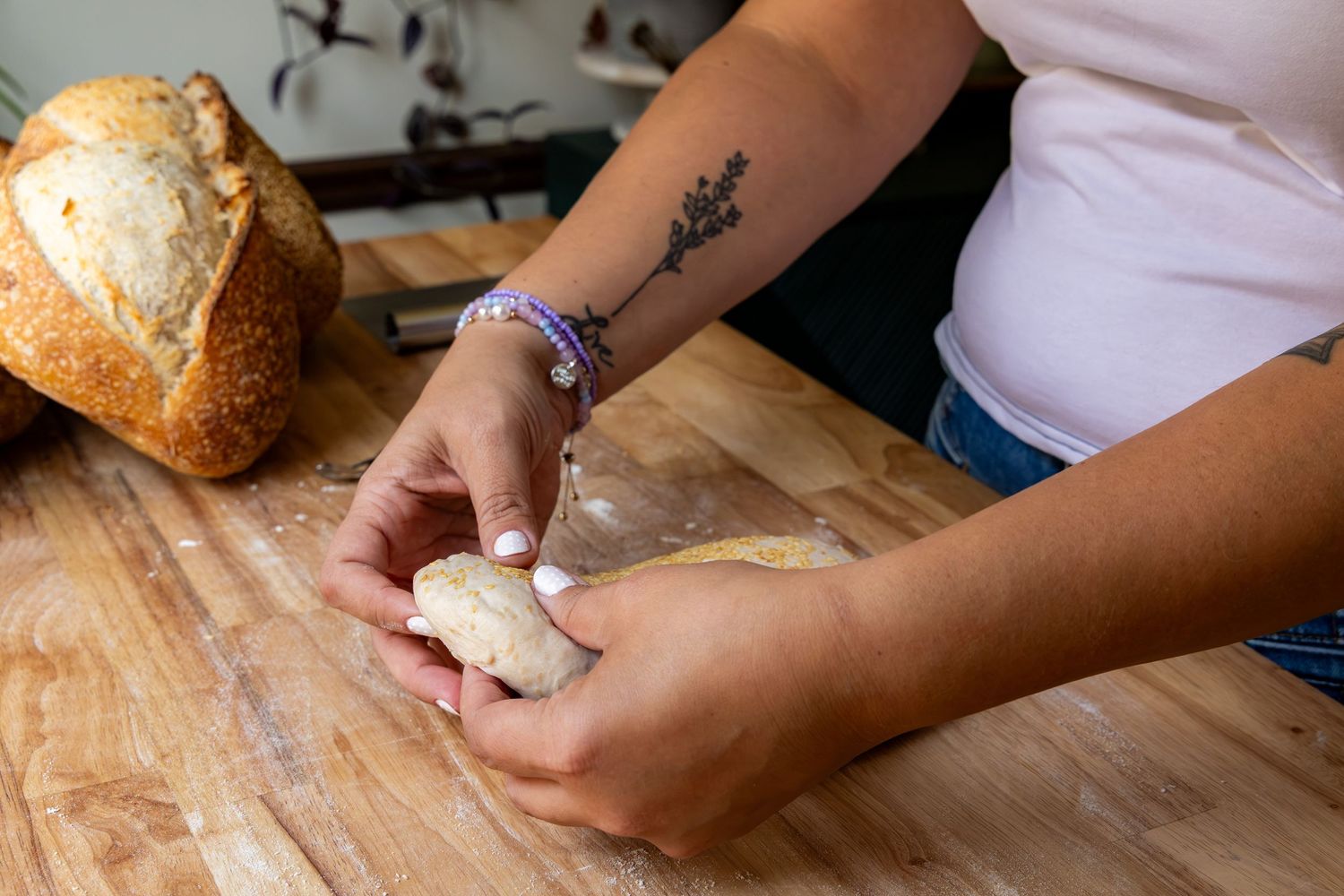 Woman with tattoo kneading bread dough on wooden cutting board with rustic sourdough loaf in background.