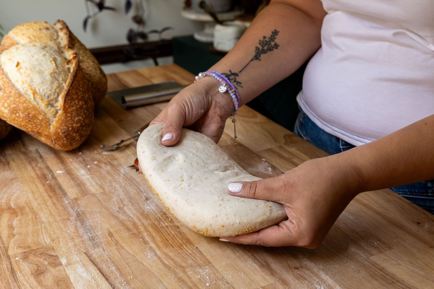 Woman shaping bread dough on a floured wooden cutting board with a baked loaf in the background.