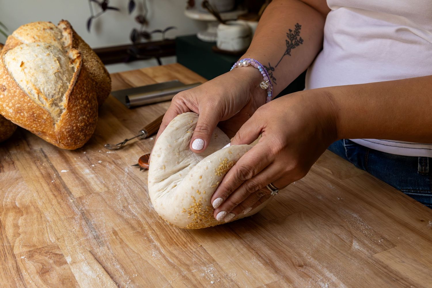 Person kneading fresh bread dough on a floured wooden cutting board with a baked loaf in the background.