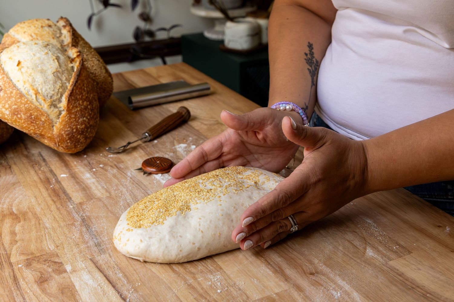 Person shaping homemade bread dough on a floured wooden cutting board with a baked loaf nearby.