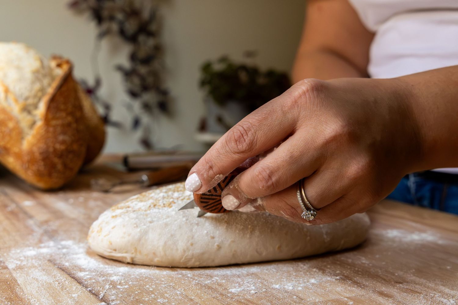 Person scoring bread dough on a floured wooden surface, with a baked loaf visible in the background.