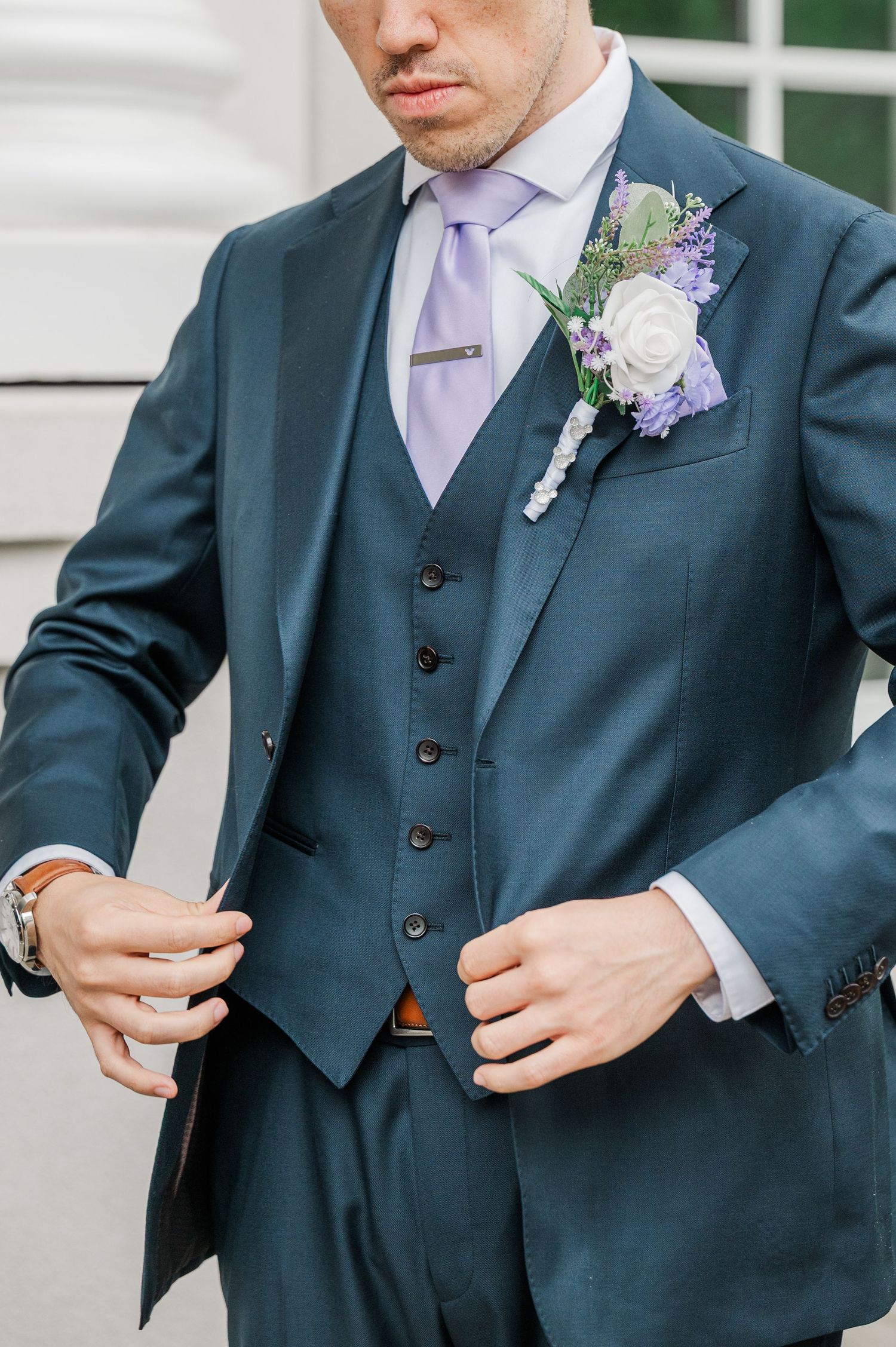 Groom in teal three-piece suit with lavender tie and floral boutonniere adjusting jacket outdoors.
