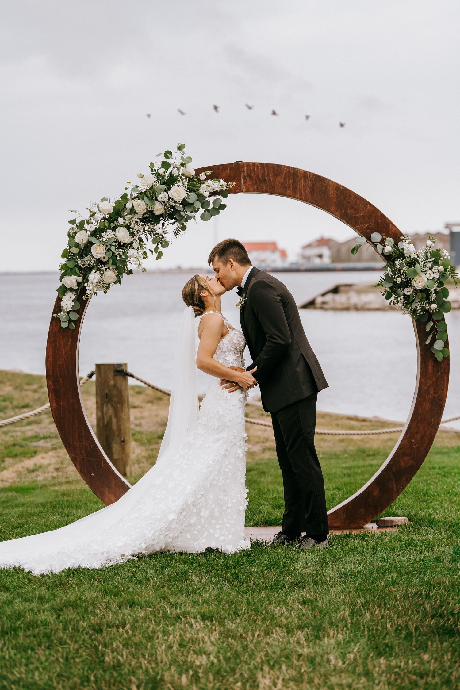 Bride and groom kissing under a circular wooden arch adorned with white flowers by the waterfront.
