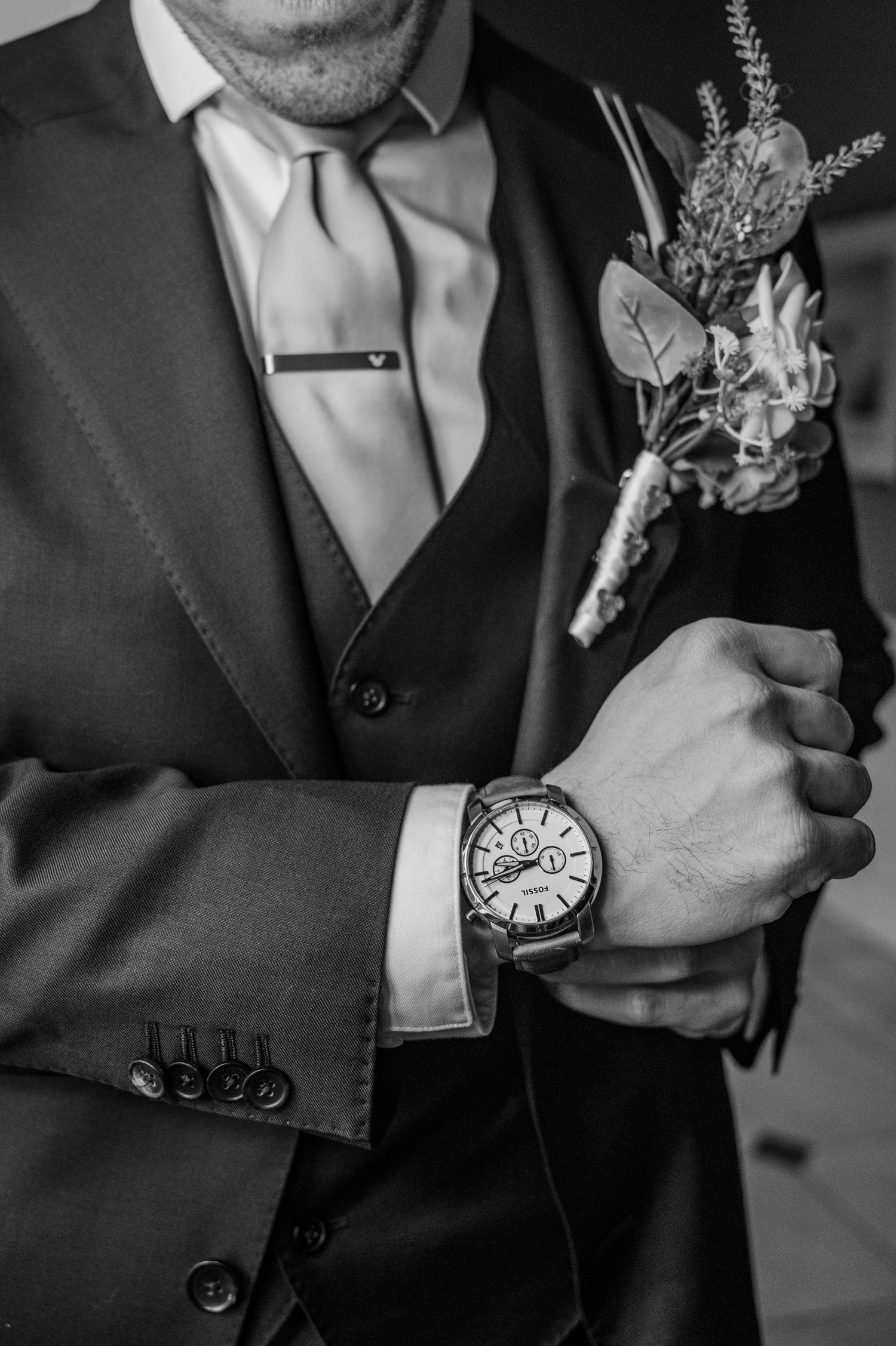 Groom in black three-piece suit with boutonniere and tie poses thoughtfully indoors in black and white wedding photo.
