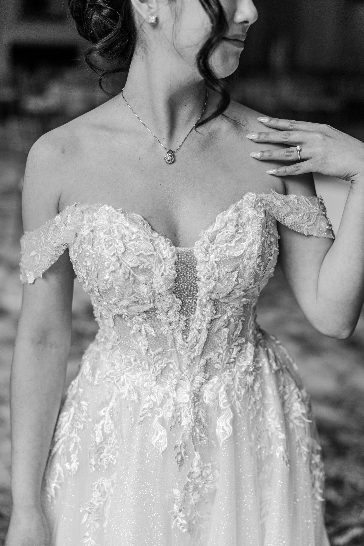 Black and white photo of a bride in an off-shoulder lace wedding gown with floral details, wearing a delicate necklace.