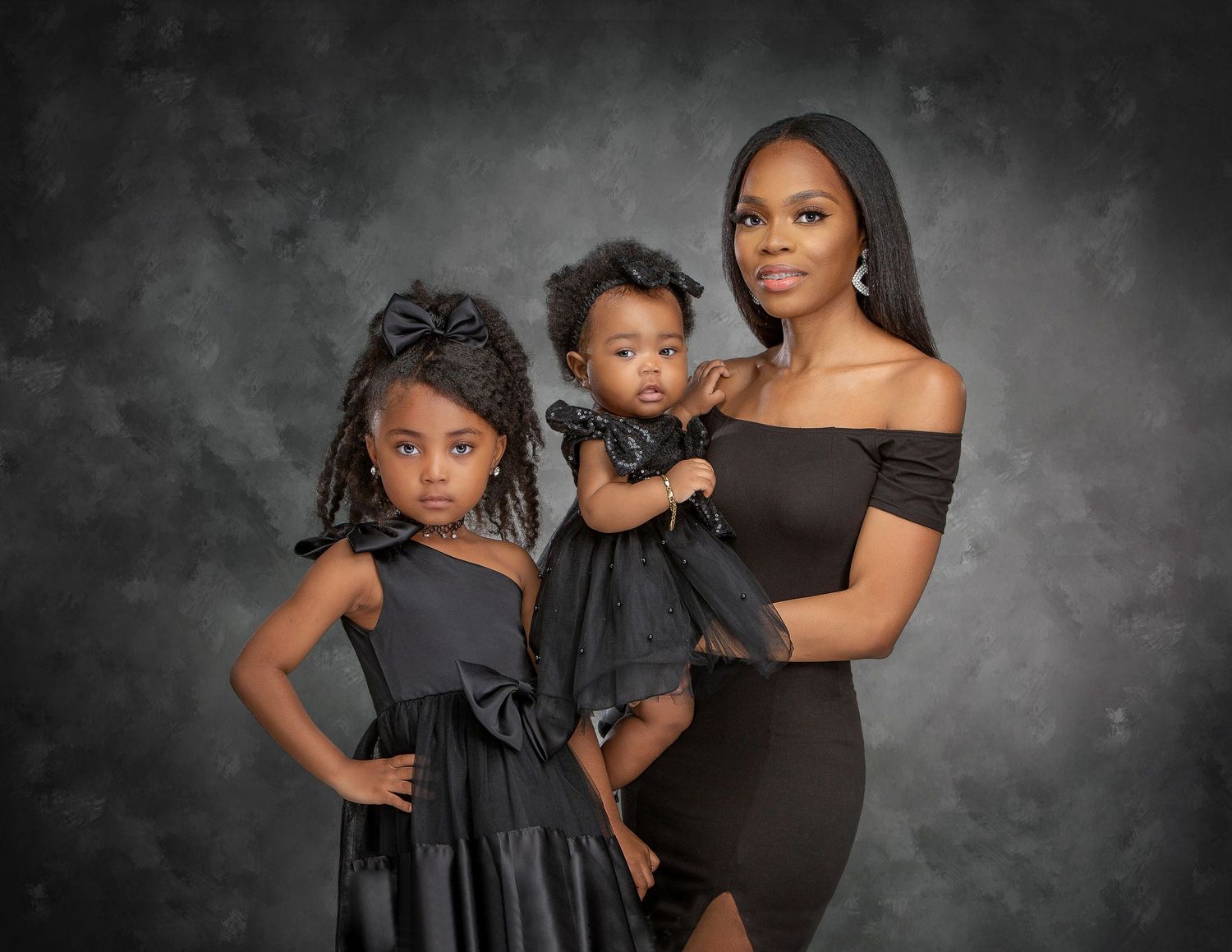 A mother in a black off-shoulder dress poses with her two young daughters, all dressed in matching black outfits.