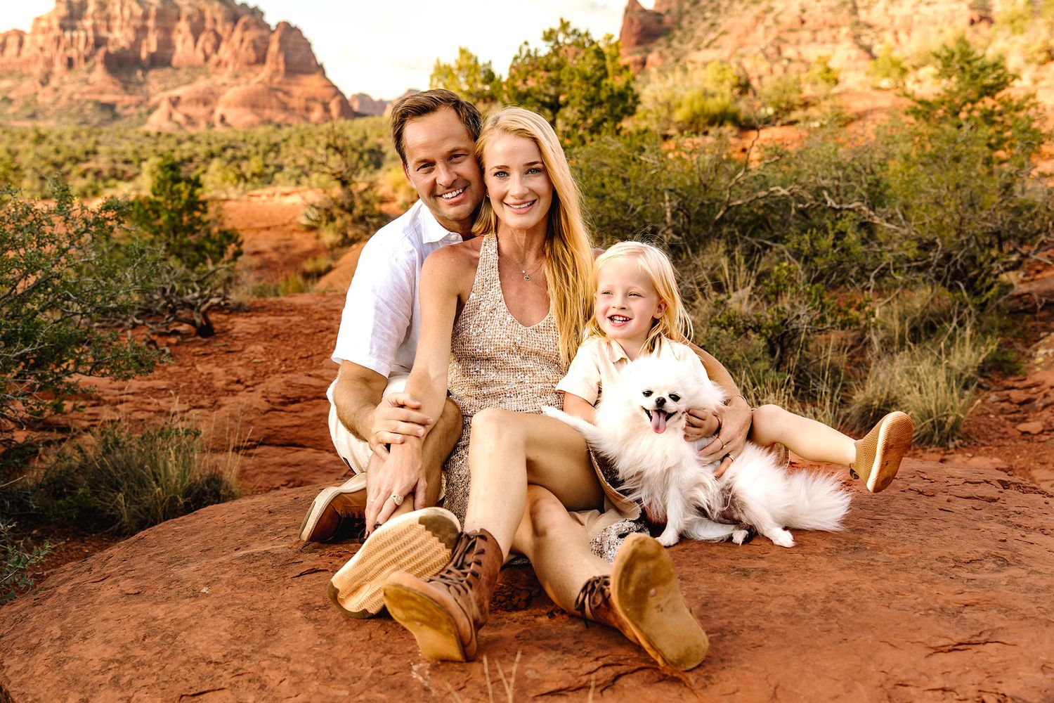 Family of three with a small dog sitting on red rocks in Sedona, Arizona, with desert landscape and red cliffs behind them.