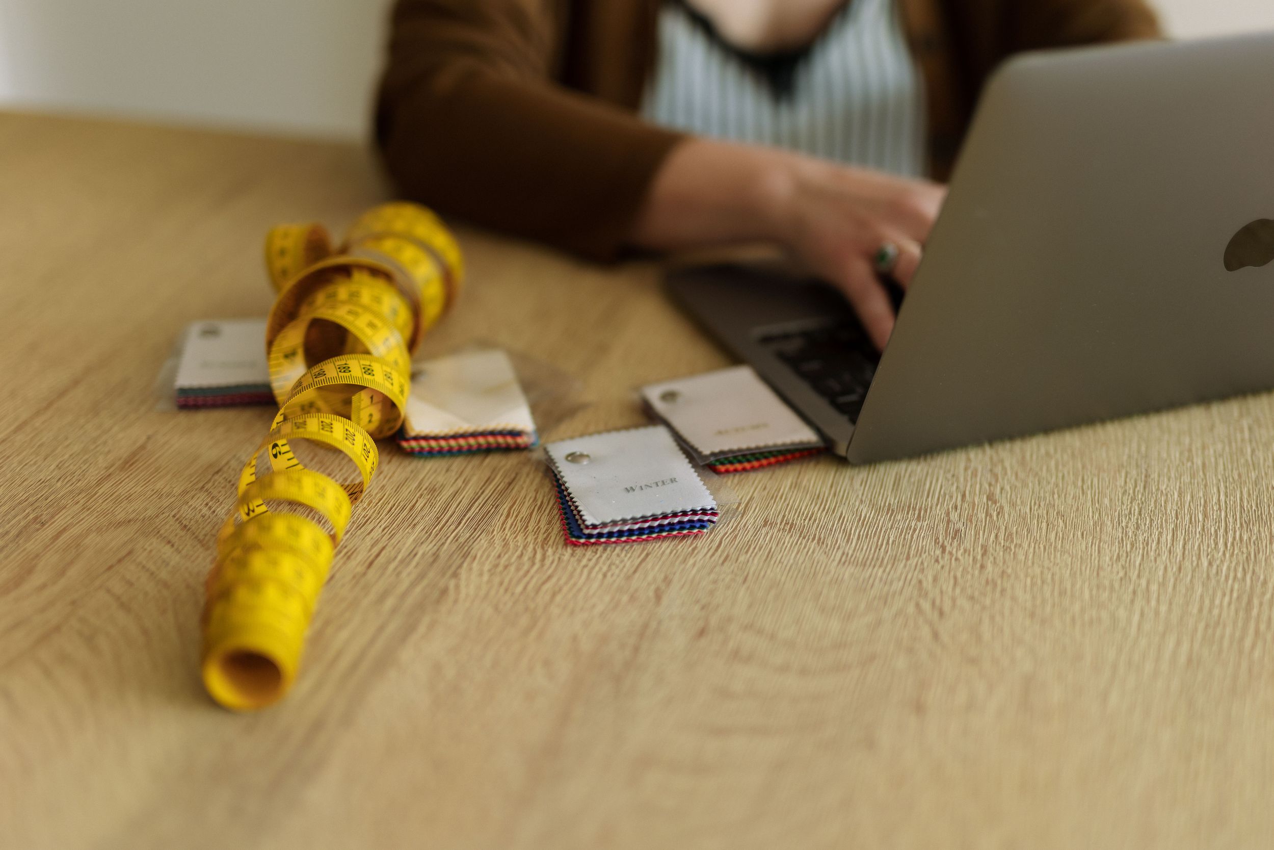 A yellow measuring tape and notebooks on a wooden desk beside a person typing on a laptop computer.