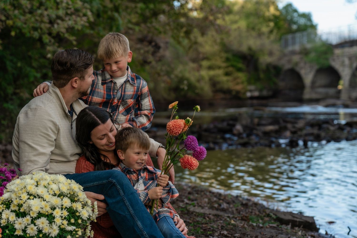 Happy family with two young children sitting by a riverside bridge surrounded by colorful autumn flowers.