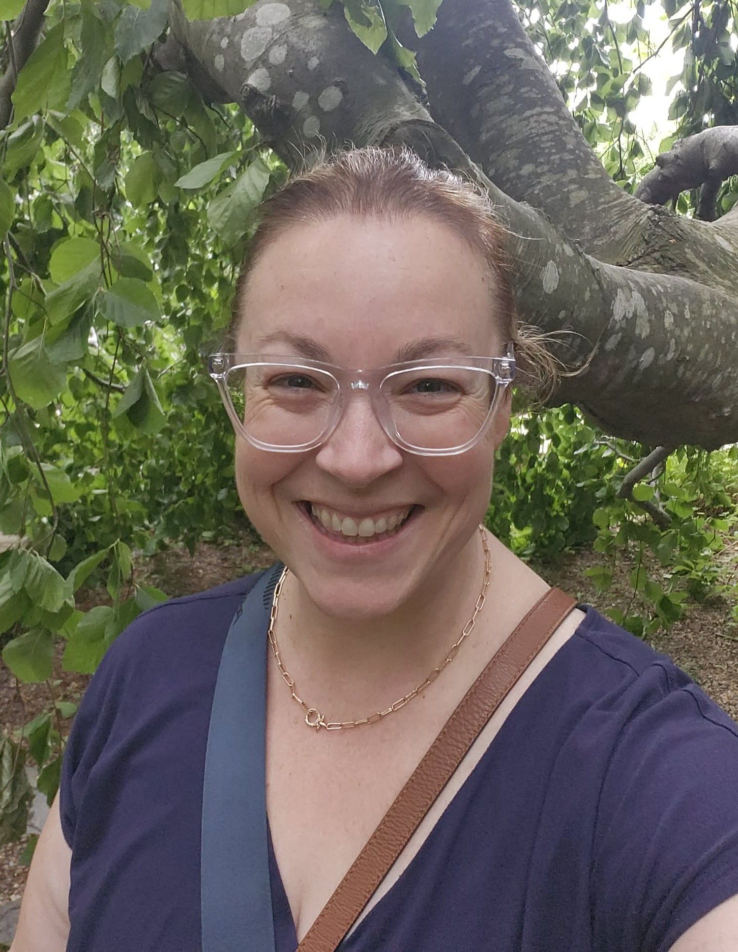 Smiling woman with red hair and clear glasses takes a selfie outdoors under a tree with lush green leaves.